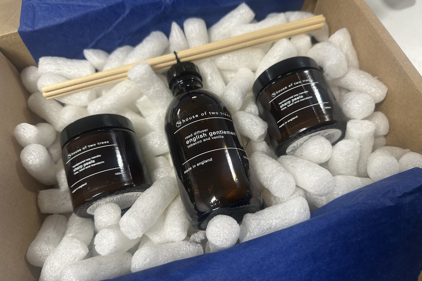 An open cardboard box with two candles and a reed diffuser packed in starch loose fill and blue tissue