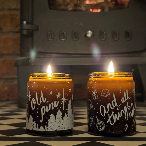 Two lit candles with decorative labels in front of a fireplace.